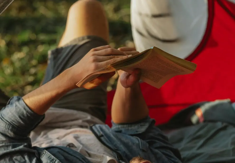 High angle of relaxed young male traveler in casual clothes lying in tent and reading interesting novel during camping in nature on sunny day
